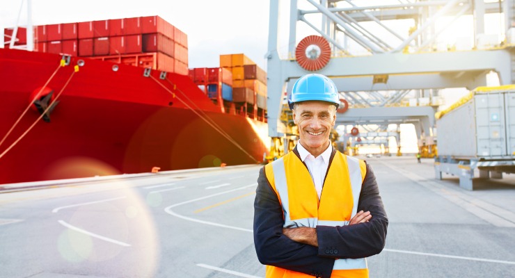 An older white male in a high visibility orange jacket and light blue hard hat, who is smiling infront of a large red sea freight container that is being loaded with red cargo containers for international shipping